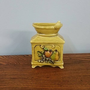 Puede incluir: Un tarro de cerámica amarillo con un diseño de veta de madera y un motivo de fruta. El tarro tiene una abertura ancha y una pequeña manija en el lateral. El motivo de la fruta incluye una pera, una manzana y uvas.