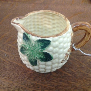 May include: A small white ceramic creamer with a brown handle and a green leaf design. The creamer is shaped like an ear of corn.