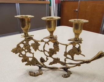 Puede incluir: Un candelero de metal dorado con un diseño de vid. El candelero tiene tres portavelas y un mango largo y curvo.
