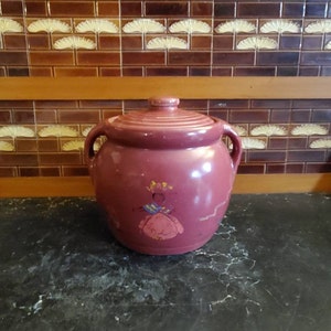Peut inclure: Un pot à biscuits en céramique rose vintage avec un motif peint d'une femme en robe rose. Le pot a deux poignées et un couvercle.