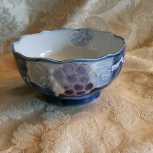 May include: A blue and white ceramic bowl with a grape design. The bowl has a scalloped rim and a footed base.