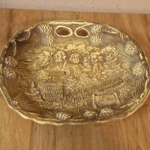 May include: A gold-colored ceramic dish with a raised design of Mount Rushmore and the names of the presidents carved into the mountain. The dish has a pine cone design around the edge.