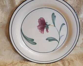 Puede incluir: Un plato de cena de cerámica blanca con un borde negro y una sola flor rosa con hojas verdes en el centro.