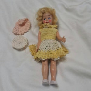 May include: A vintage doll with blonde hair and blue eyes wearing a yellow and white crocheted dress and white shoes. The doll is holding a pink crocheted hat and a white crocheted hat.