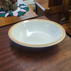 May include: A white ceramic bowl with a light brown rim. The bowl is empty and sitting on a wooden surface.