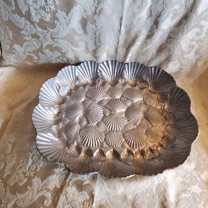 May include: A silver oval serving tray with a raised design of seashells. The tray is made of metal and has a shiny finish.