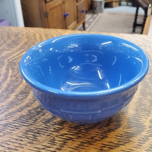 May include: A vibrant blue ceramic bowl with a glossy finish. The bowl has a rounded shape with a textured, basketweave-like pattern around the exterior. The bowl is sitting on a wooden table.
