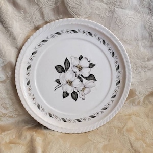 May include: White ceramic plate with a floral design featuring white flowers with yellow centres and green leaves. The plate has a scalloped edge and a grey and white border.