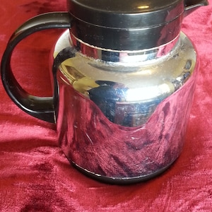 May include: A silver and black thermal carafe with a black lid and a handle. The carafe has a shiny, reflective surface.