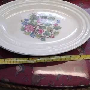 Puede incluir: Un plato ovalado blanco con un diseño floral en tonos de rosa, azul y verde. El plato tiene un borde plateado y mide aproximadamente 30 cm de largo.