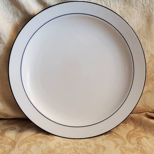 May include: A white ceramic plate with a black rim and a thin blue accent line. The plate is round and has a simple, elegant design, suitable for serving food. The plate is likely 10-12 inches in diameter.