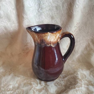 May include: A brown ceramic pitcher with a light brown glaze around the top. The pitcher has a rounded body and a handle.
