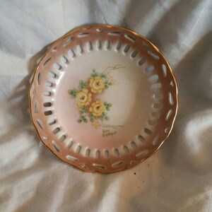 May include: A small, round, white porcelain dish with a pink rim and a gold trim. The dish has a cut-out design around the edge and features a hand-painted floral design with yellow roses. The dish has the words "Sue Lightfoot" written on the bottom.