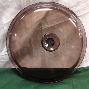 Op de afbeelding: Een doorzichtig glazen deksel met een donkerbruine tint. Het deksel heeft een knop in het midden.