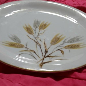 May include: White oval ceramic plate with a gold rim and a design of wheat stalks in shades of brown and gray.