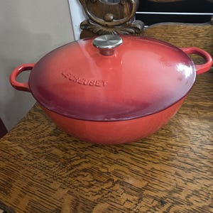 Peut inclure: Une cocotte Le Creuset en fonte émaillée rouge avec un couvercle et des poignées latérales. Le pot a un bouton argenté sur le couvercle. La cocotte est rouge vif et est posée sur une surface en bois.
