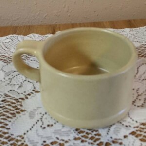 Puede incluir: Una taza de cerámica de color crema con un asa redondeada. La taza está vacía y se encuentra sobre un encaje blanco.