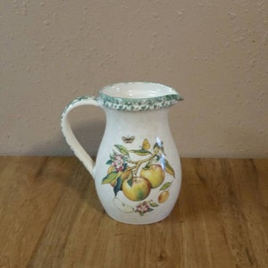 Puede incluir: Jarra de cerámica blanca con borde verde y un diseño floral con manzanas, hojas y mariposas.