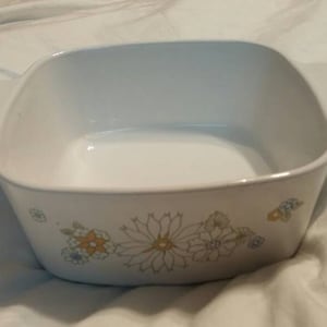 Puede incluir: Plato para hornear de cerámica blanca con un diseño floral en tonos amarillos, azules y verdes. El plato tiene dos asas a los lados.