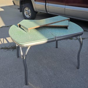 May include: A green laminate table with a chrome metal edge and four curved chrome legs. The tabletop features a mottled pattern and includes two removable rectangular extension leaves stacked on top of the main surface, showing exposed wood edges.