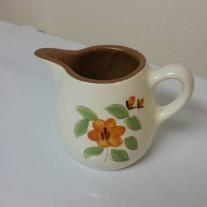 Puede incluir: Un pequeño creamer de cerámica blanca con un borde marrón y un diseño floral con flores naranjas y hojas verdes.