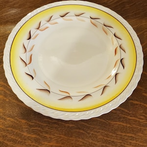 May include: A white ceramic bowl with a yellow rim and brown leaf design. The bowl has a scalloped edge.