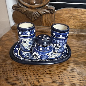 Könnte beinhalten: Ein Keramik-Salz- und Pfeffer-Set mit passendem Tablett. Das Set hat ein blau-weißes Blumenmuster mit dunkelblauem Rand. Es enthält zwei zylindrische Behälter und einen runden Shaker, die alle auf einem ovalen Tablett ruhen.
