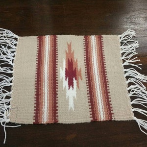 May include: A beige woven placemat with red and white stripes and a diamond pattern in the center. The placemat has white fringe on all sides.