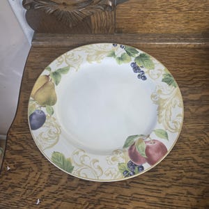 May include: A white ceramic plate with a gold rim, decorated with fruit illustrations. The design includes pears, plums, apples, and grapes, complemented by gold scrollwork. The plate is displayed on a wooden surface.