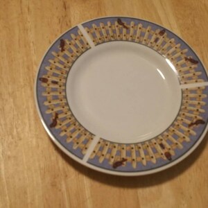 May include: A white ceramic plate with a blue and brown border design. The border features a repeating pattern of a picket fence with birds.
