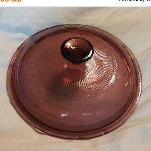 Op de afbeelding: Een doorzichtig glazen deksel met een paarse tint en een knop bovenop. Het deksel heeft een getextureerd, geribbeld patroon.