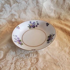 May include: A white ceramic bowl with a gold rim and a floral design featuring purple and yellow flowers. The bowl is on a white tablecloth with a floral pattern.