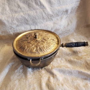 May include: A vintage brass pot with a lid featuring an intricate design of figures. The pot has a wooden handle and a brass loop handle.