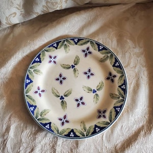 May include: A white ceramic plate with a blue and green floral design. The plate has a scalloped edge and is decorated with small blue and green flowers.