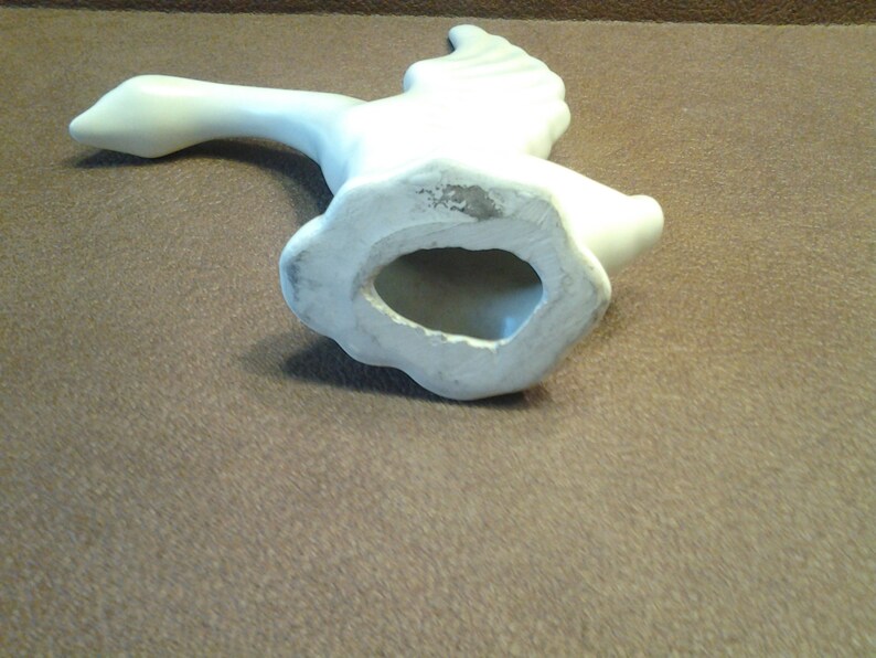 May include: A white ceramic goose figurine in mid-flight, with outstretched wings and a long neck. The underside reveals a hollow base. The sculpture is smooth and appears to be unpainted, with a matte finish.