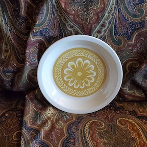 Może przedstawiać: Biała ceramiczna miska z żółtym i białym wzorem kwiatowym. Miska ma falisty brzeg i jest ozdobiona stylizowanym kwiatem w środku.