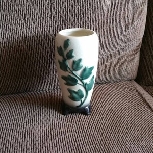 May include: A white ceramic vase with a green leafy design. The vase has three small feet and is sitting on a brown fabric surface.