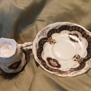 May include: A white teacup and saucer set with black and gold floral designs. The saucer has a scalloped edge.