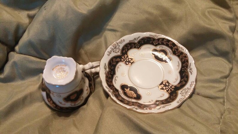 May include: A white teacup and saucer set with black and gold floral designs. The saucer has a scalloped edge.