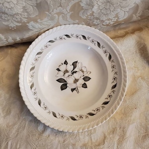 May include: White soup bowl with a floral design of white dogwood blossoms with green leaves and stems. The bowl has a white background with a brown and green border.