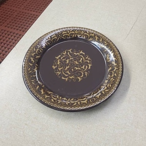 Op de afbeelding: Een bruin keramiek bord met een gouden decoratief ontwerp rond de rand en een gouden bloemenpatroon in het midden.