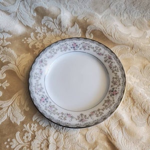 Può includere: Un piatto in porcellana bianca con un design floreale argento e grigio. Il piatto ha un bordo ondulato e un bordo rialzato.