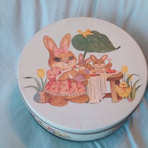 以下が含まれることがあります： 白い背景に3羽のウサギが描かれた、ヴィンテージの丸いブリキのクッキー缶。一番大きなウサギはピンクのドレスを着て、緑の傘を持っています。他の2羽のウサギは白いテーブルクロスと黄色いひよこがいるテーブルに座っています。