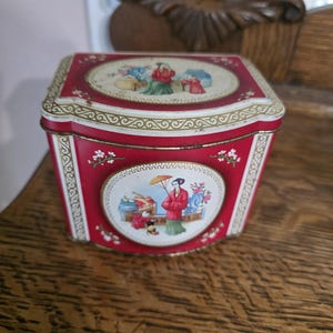 Op de afbeelding: Een rode, vintage blikken doos met een scharnierend deksel. De doos heeft ovale illustraties van mensen in traditionele kleding, met bloemen en gouden accenten. De doos staat op een houten oppervlak.