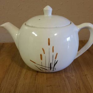 May include: White ceramic teapot with a lid and a brown and black abstract design of tall grass.