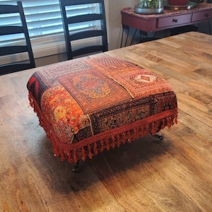 May include: A square ottoman with a patterned, tapestry-like top in shades of red, orange, and black. The ottoman has a red beaded fringe and sits on short, dark metal legs. The piece is on a wooden surface.