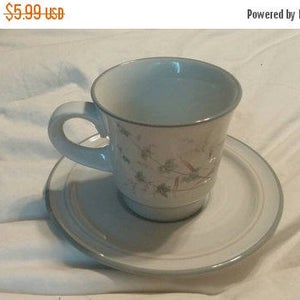 May include: A white teacup and saucer set with a light blue trim. The cup has a handle and a floral design. The saucer is round and matches the cup's trim. The set is on a white surface.