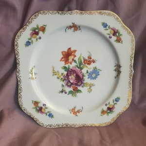 Può includere: Un piatto da portata quadrato bianco con un bordo dorato e un motivo floreale in tonalità di rosa, arancio, giallo e blu.