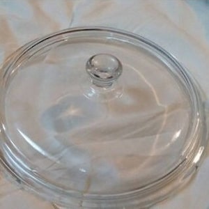 Op de afbeelding: Een doorzichtig glazen deksel met een knop aan de bovenkant. Het deksel is rond en heeft een licht verhoogde rand.