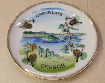 Puede incluir: Un plato de cerámica blanco con borde dorado presenta una ilustración colorida del lago Crater, en Oregón. La imagen incluye un lago, una isla llamada "Wizard Island" y pinos con piñas. Los textos "Crater Lake" y "Oregon" están impresos en negro.